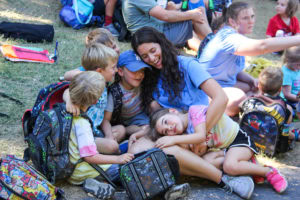 A group of children sits closely around a smiling counselor outdoors, some leaning on her, with backpacks and other people visible in the background.
