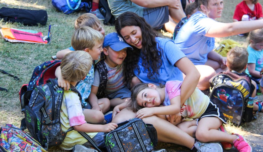 A group of children sits closely around a smiling counselor outdoors, some leaning on her, with backpacks and other people visible in the background.