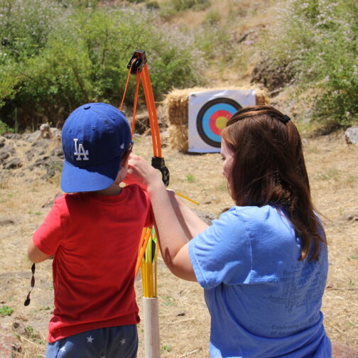 An adult helps a child aim a bow and arrow at an archery target outdoors on a sunny day.