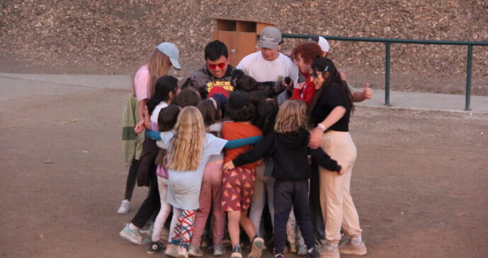 A group of children and adults stand in a circle outdoors, hugging each other on a dirt surface near a wooden backdrop and metal railing.