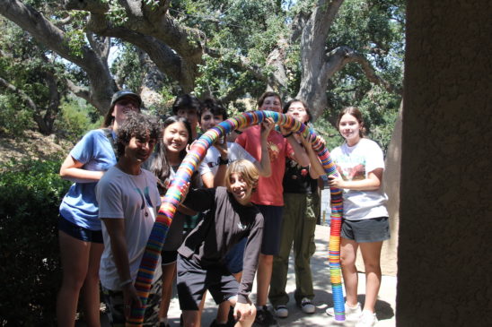 A group of smiling teenagers stand outdoors under trees, holding a long, colorful object made of stacked plastic disks.
