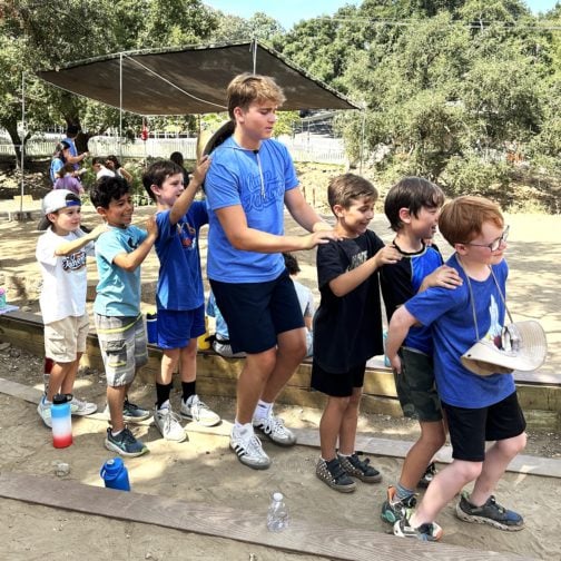 A group of boys stand outdoors in a line, each with hands on the shoulders of the person in front of them, forming a conga line. They appear to be at a camp or playground.