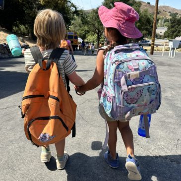 Two young children with backpacks hold hands while walking on a paved road toward a parked school bus on a sunny day.