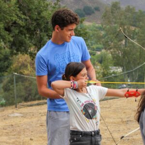 A teenage boy helps a young girl aim a bow and arrow outdoors, with trees and hills in the background.