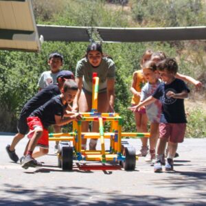 A group of children push and guide a colorful, wheeled structure outdoors, supervised by an adult.
