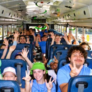 Children and counselors sitting on a school bus smiling and making peace signs, with green and yellow decorations hanging from the ceiling.