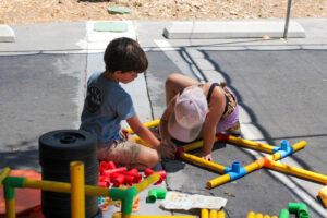 Two children are sitting on pavement assembling colorful tubes and connectors, with various construction toys scattered around them.