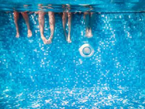 Four Summer Camp Children’s legs and feet are visible underwater as they sit on the edge of a swimming pool with blue mosaic tiles.