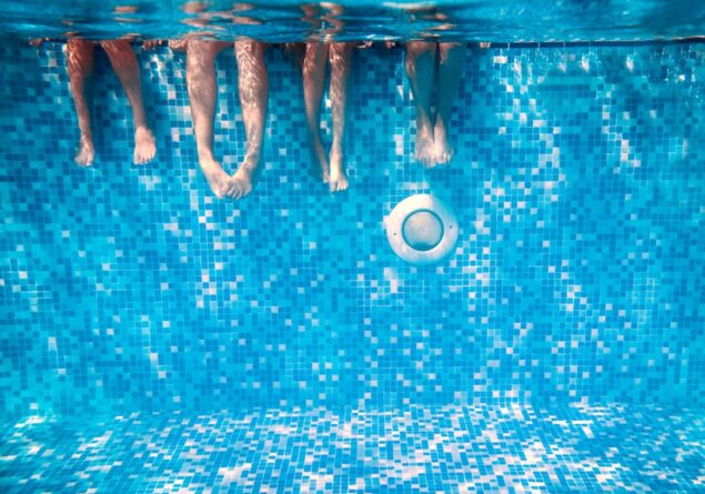 Four Summer Camp Children’s legs and feet are visible underwater as they sit on the edge of a swimming pool with blue mosaic tiles.