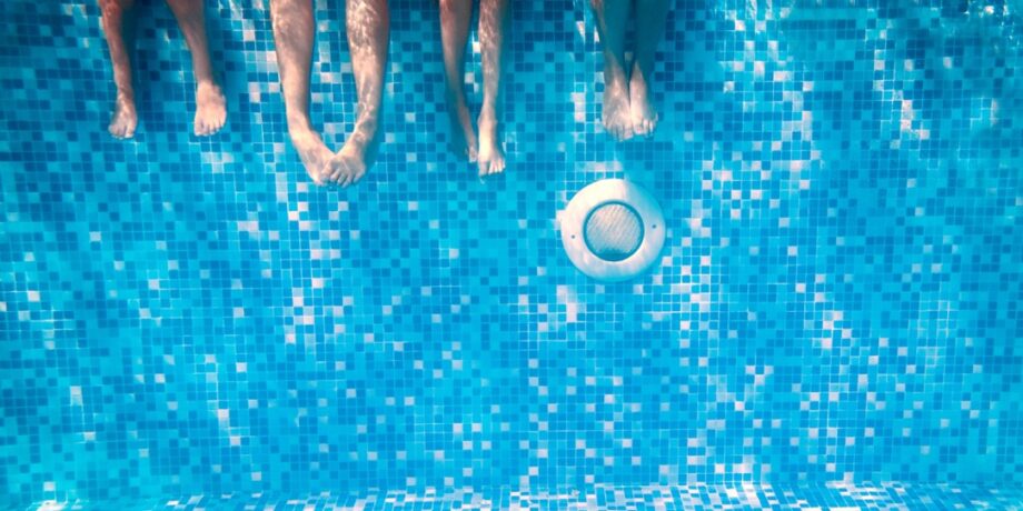 Four Summer Camp Children’s legs and feet are visible underwater as they sit on the edge of a swimming pool with blue mosaic tiles.