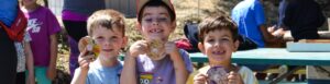 Three young boys stand outdoors, smiling and holding up decorated wooden crafts while other people sit and work at a picnic table in the background.