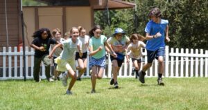 A group of children and one adult run together on grass near a white picket fence, with a building and trees in the background.