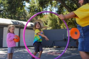 A child runs through a large hula hoop held by an adult while another child waits nearby holding a plastic disc in an outdoor area.