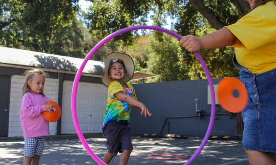 A child runs through a large hula hoop held by an adult while another child waits nearby holding a plastic disc in an outdoor area.