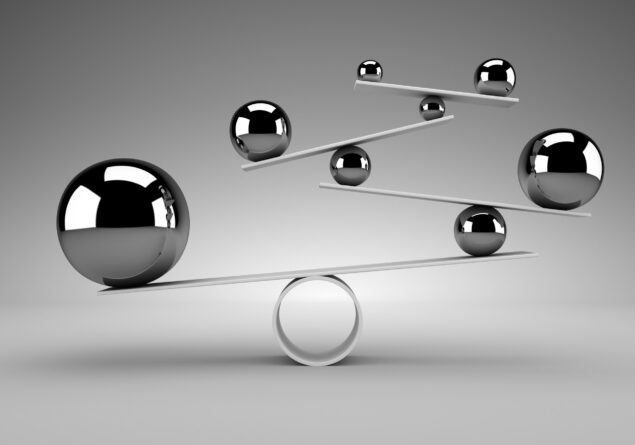 Six metallic spheres of varying sizes are balanced on white flat rods and a circular base, creating a geometric structure against a neutral gray background.