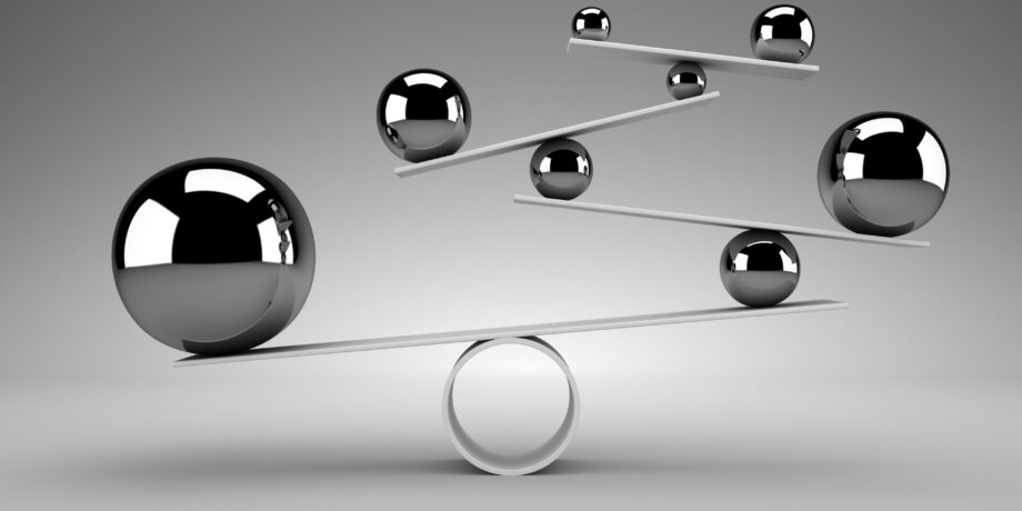 Six metallic spheres of varying sizes are balanced on white flat rods and a circular base, creating a geometric structure against a neutral gray background.