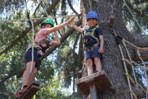 Two children in safety gear and helmets high-five on a ropes course in a forest, standing on wooden platforms surrounded by trees and ropes.